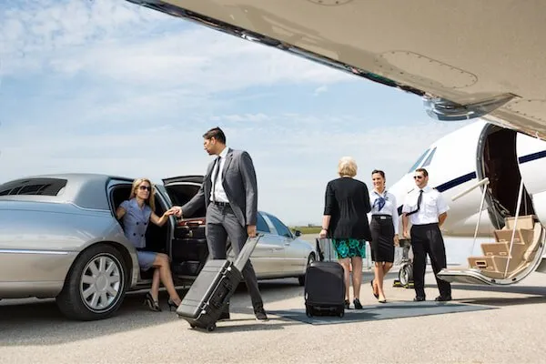 airport-car-service-branford-1920w A woman exits a car as a man and two others stand near a private jet, one holding luggage. Two crew members are positioned close to the jet's stairs.