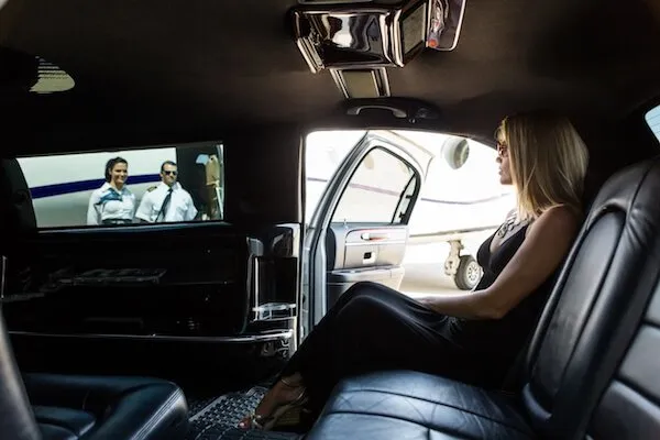 airport-transfers-1920w Person in a black dress sits in a car, watching a movie on a screen. An airplane is visible outside through the open door.