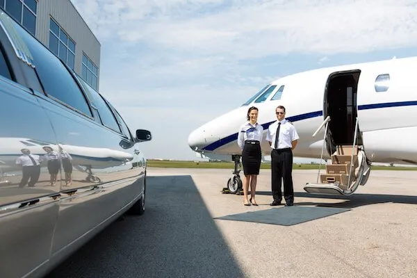 best-JFK-airport-transportation-service-1920w Two pilots stand beside a private jet on an airport tarmac, with a limousine parked nearby under a clear sky.