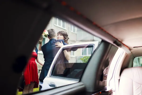 west-haven-wedding-limousine-rental-1920w View from inside a car showing a person in a formal blue outfit embracing a person in a blue dress near an open door. Another person in a red dress stands nearby.